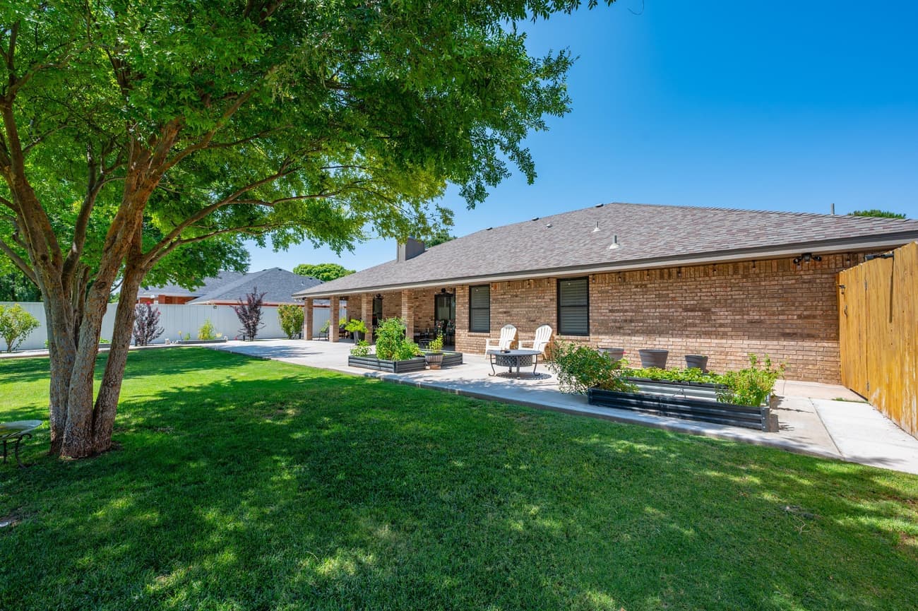The back of the house from under a shade tree: a long covered patio, raised metal planting beds, a wide lawn and a solid privacy fence.
