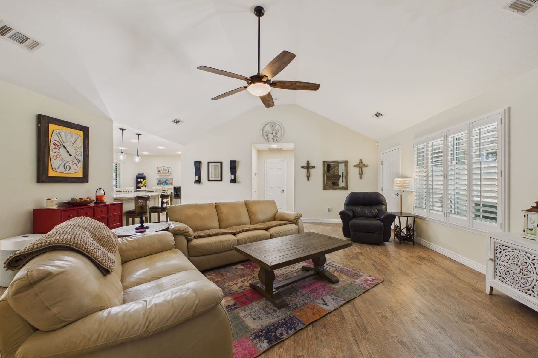 The living room: a vaulted ceiling with a fan, two large sofas and a recliner around a wooden coffee table, plantation shutters, and a red painted cabinet against a cream wall.